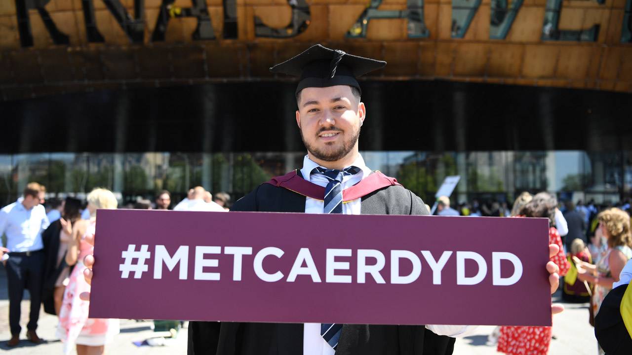 Student holding Welsh hashtag sign at graduation in front of WMC
