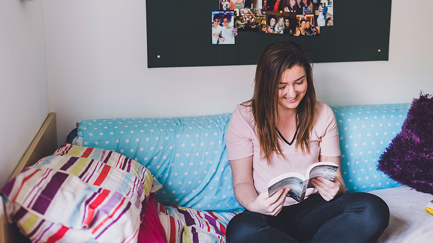 Female student sat on a bed in her student accommodation reading a book