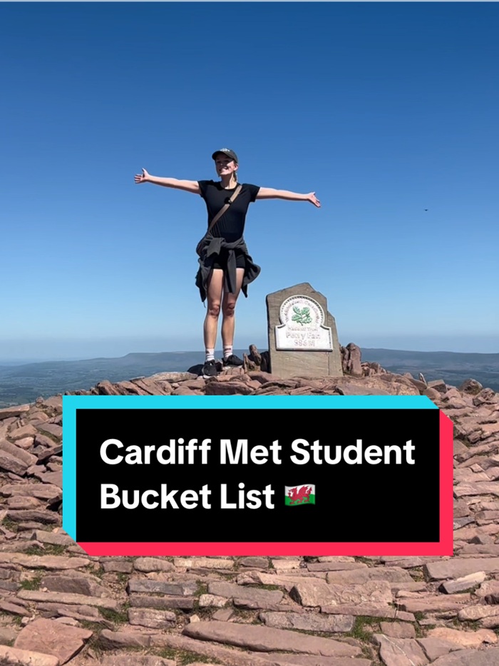 A young woman stands with arms outstretched at the peak of a mountain on a sunny day