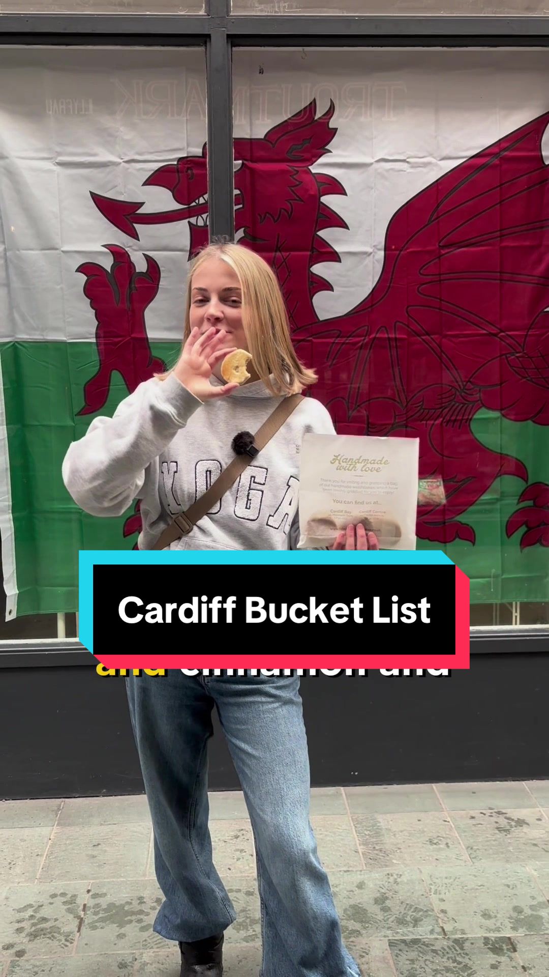 A young woman eats a Welsh cake in front of a large Welsh flag