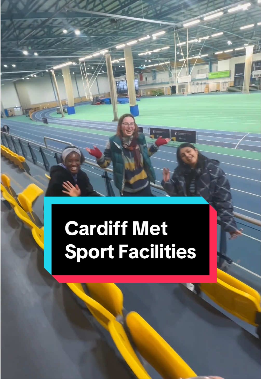 Three students pose for a photograph while sitting or standing in the spectator seats of the National Indoor Athletics Centre