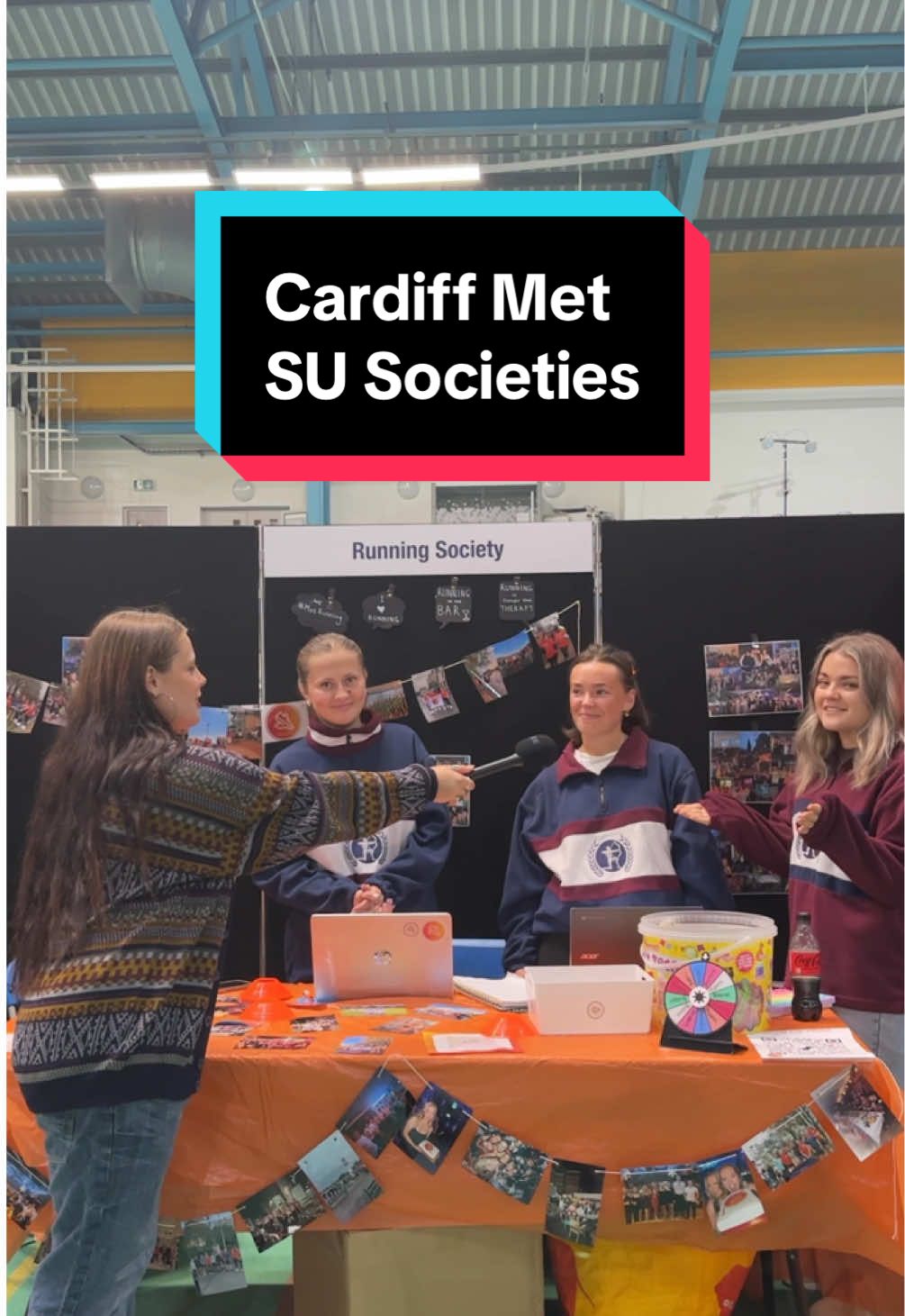 Three students hosting the Running Society stand at the Freshers' Fayre are asked a questions by a young woman holding a microphone