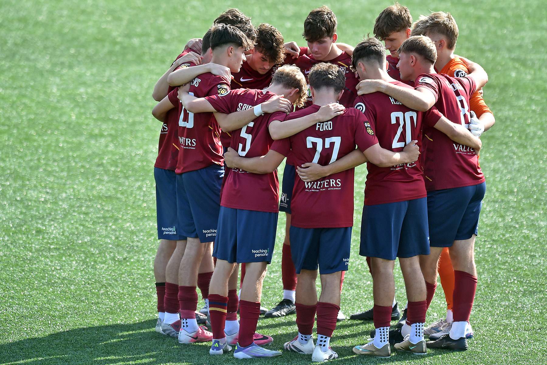 A group of young men huddled together on a football pitch. They are wearing maroon shirts and dark blue shorts.