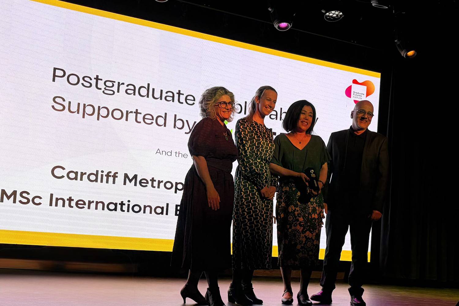 A group of four people pose for a photo in front of large display screen. One of them is holding an award in their hands.