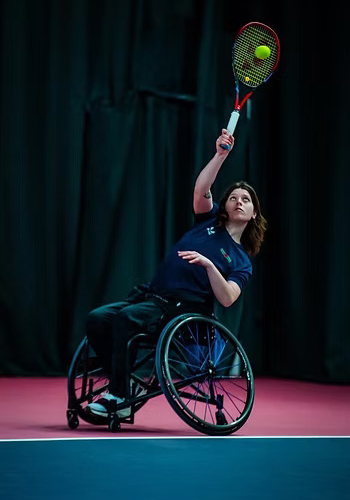 A female wheelchair tennis player on an indoor court serving a ball.
