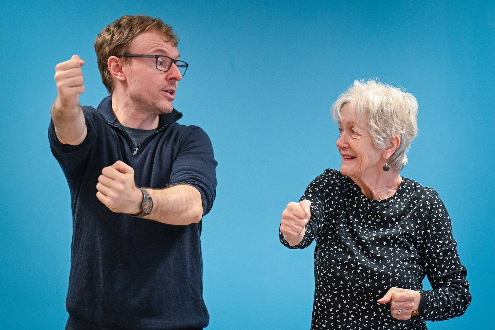 A man demonstrates a martial arts movement standing next to an older woman. The woman is copying the movement.