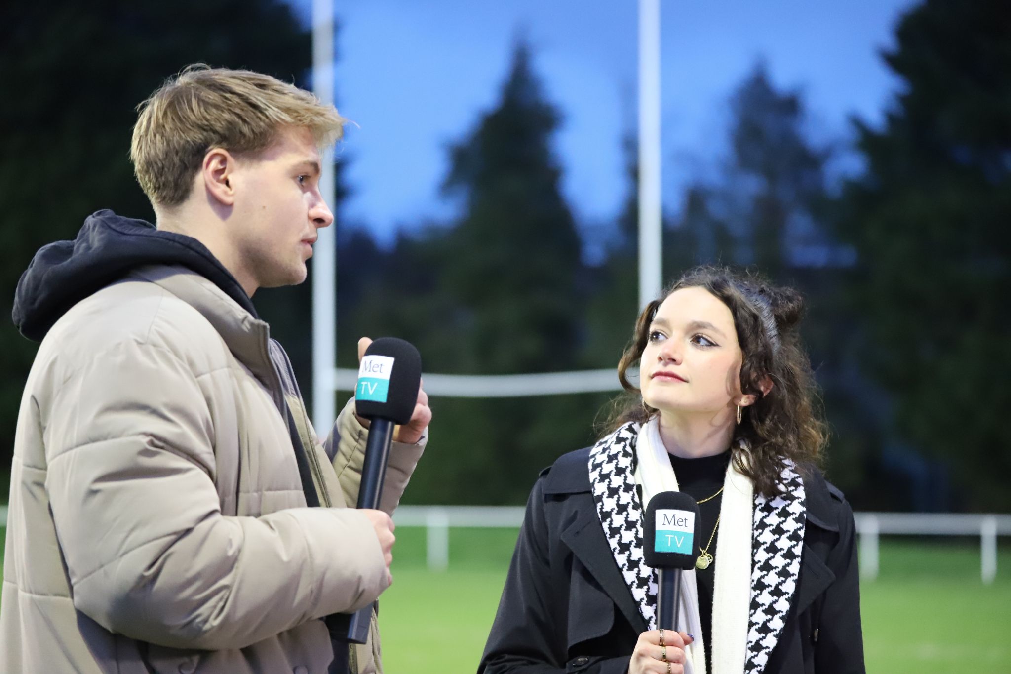 Two people stand on a rugby pitch. They are holding microphones with Met TV printed on them.