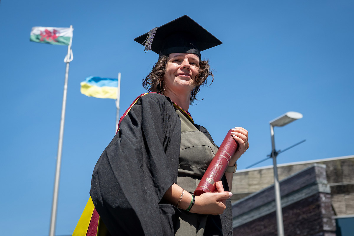 A woman wearing a graduation cap and gown pictured against a bright blue sky. In the background are flag poles with the flags of Wales and Ukraine.