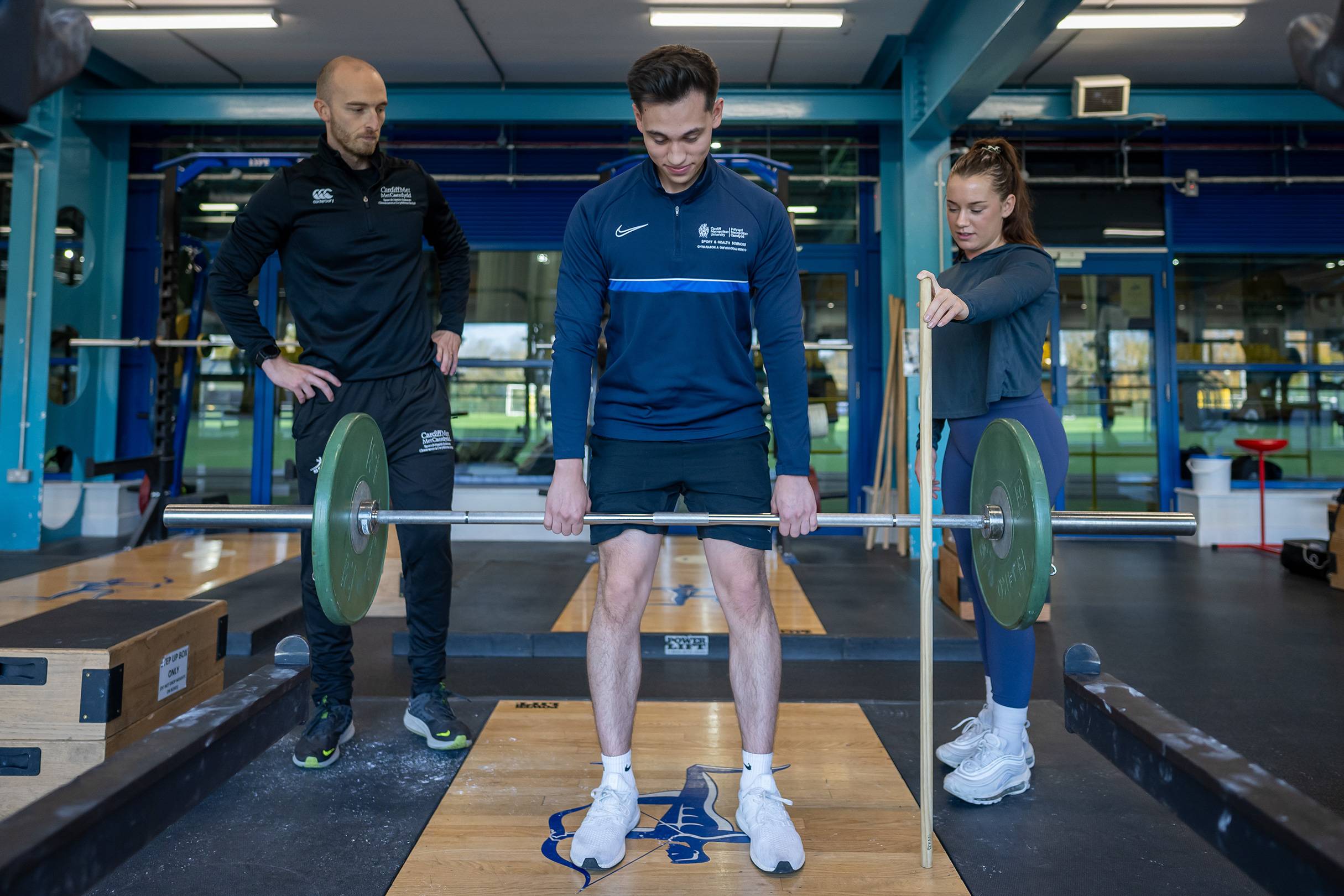 A person lifts a weight in a gym while two other people watch.