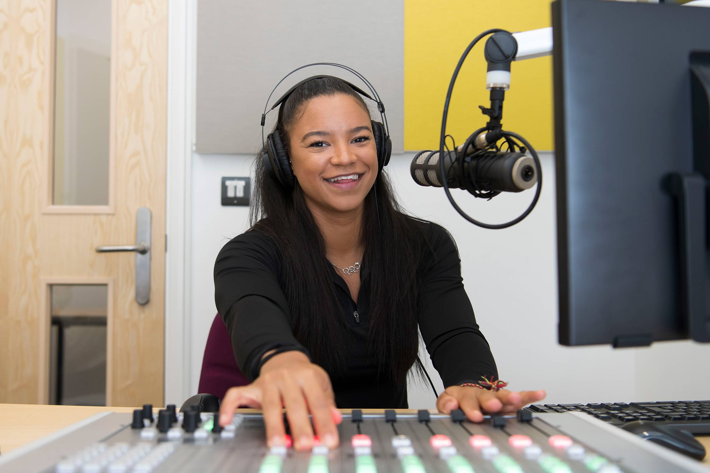 A person wearing headphones sits at a sound mixing desk. In front of them is a microphone mounted on an adjustable arm.