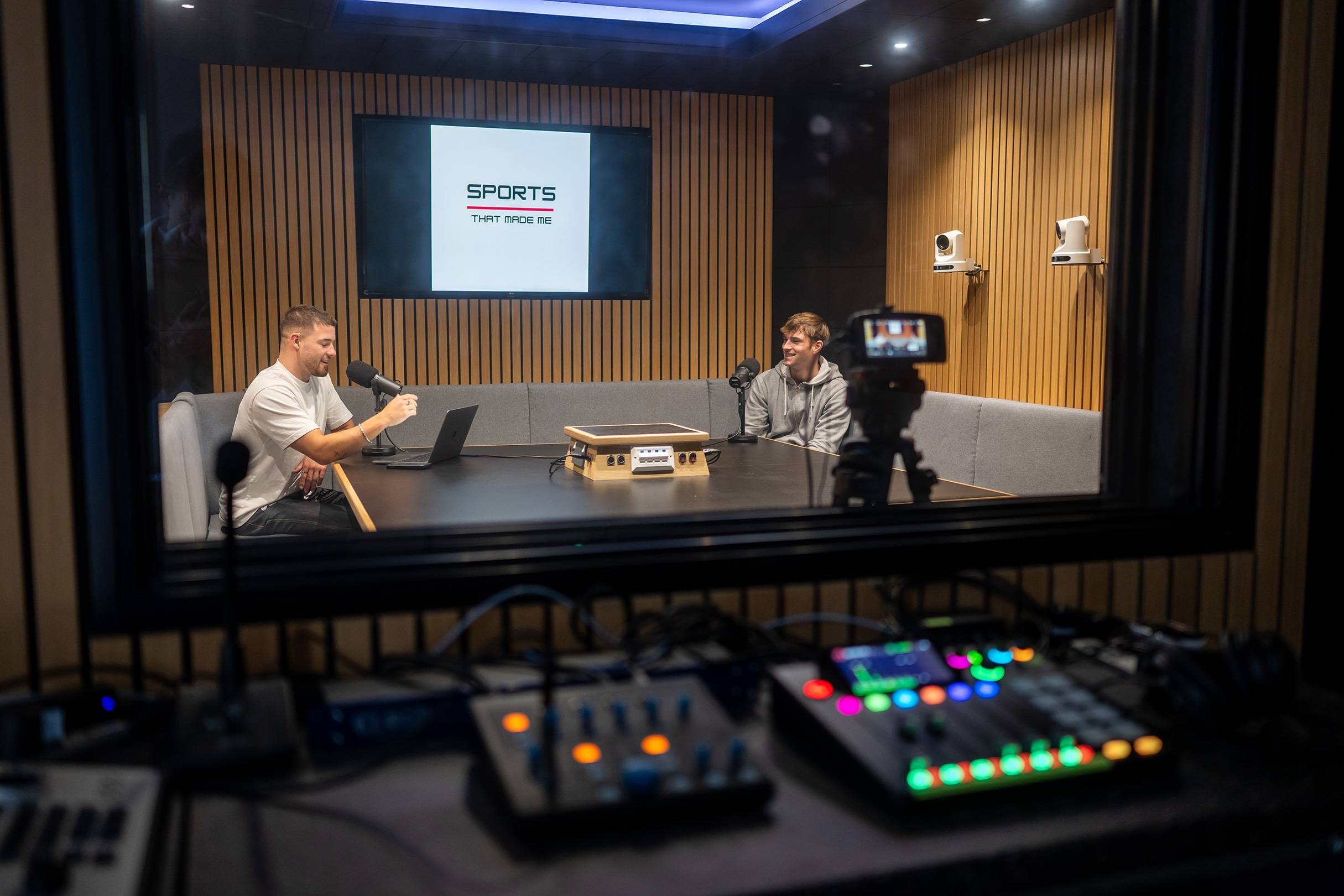 Two people sit at a table in a sound booth talking to one another. In front of them are microphones and other audio equipment.