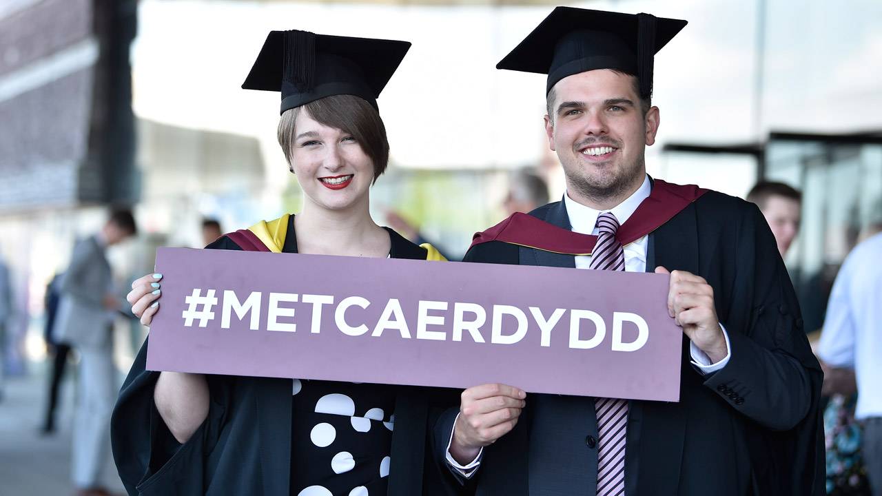 Two students holding Welsh hashtag sign at graduation